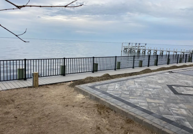 Riverside walkway with black railing, paved patio, and a calm body of water under a cloudy sky