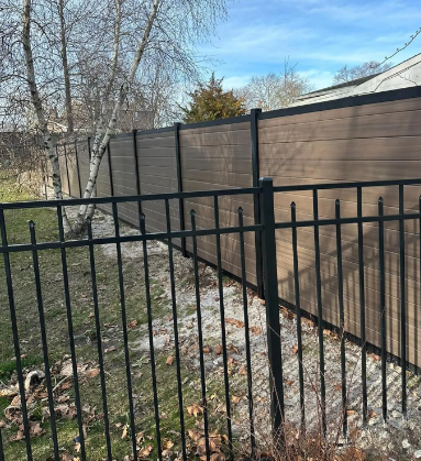 Black metal fence in front of a brown wooden privacy fence beside a grassy yard with trees and a house.