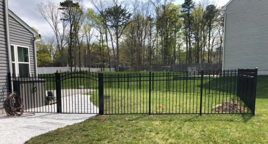 Black metal gate between two gray-sided houses opening to a grassy yard and wooded background