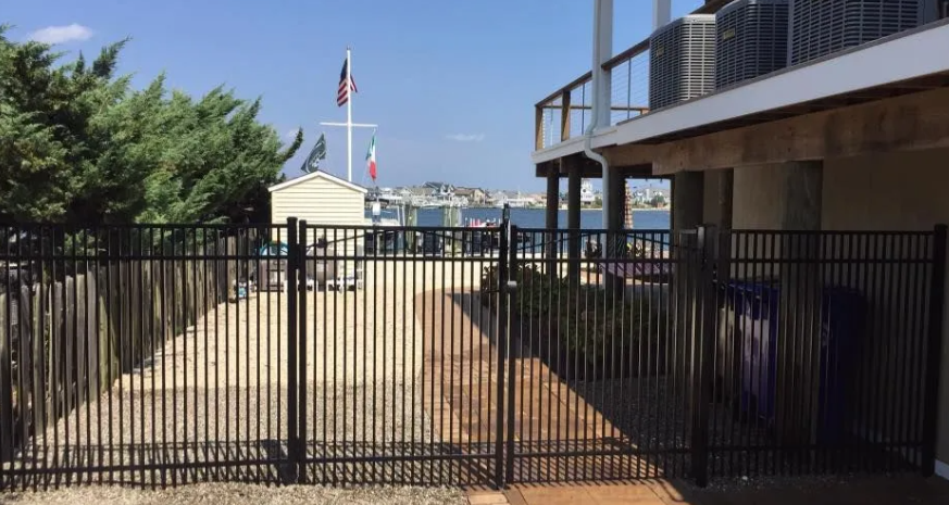 Black metal gate opening to a paved courtyard with a building, flags, and trees in bright sunlight