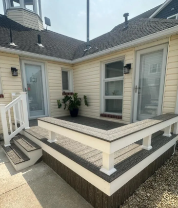 Small raised wooden porch with white railings, steps, potted plant, and beige house siding with windows and shutters
