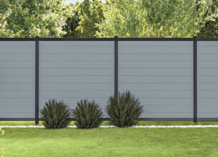 Gray privacy fence with dark posts and green shrubs in front, bordered by a grassy lawn and trees behind.