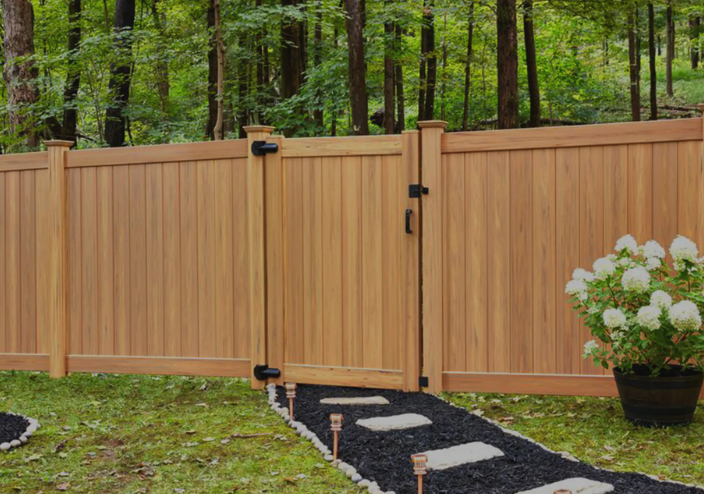 Wooden driveway gate beside a black gravel path, with potted white flowers and a wooded backdrop.