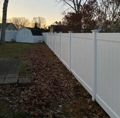 White vinyl fence along a leaf-covered yard at dusk, with sheds and bare trees in the background
