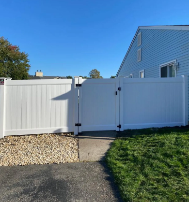 White vinyl backyard gate beside a blue house, with gravel, pavement, and green grass under a clear sky