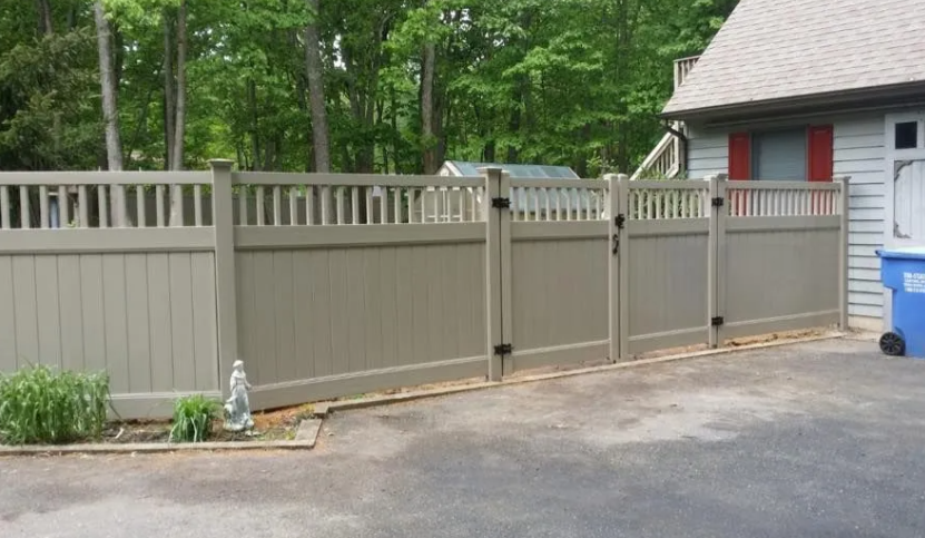 Tan fence beside a driveway and house, with trees and blue recycling bins in the background