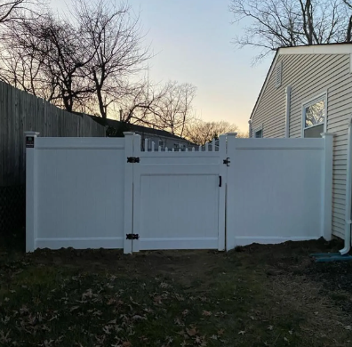 White vinyl fence and gate in a backyard at dusk, with a house on the right and bare trees behind.
