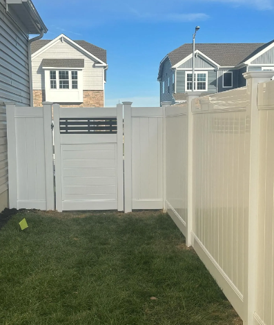Small fenced backyard with green grass and white vinyl fencing, gate, and neighboring houses in the background