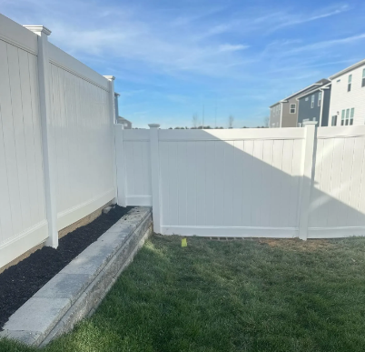 White vinyl fence corner with a concrete border and green grass under a blue sky.