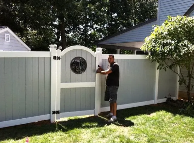 Man opening a white gate in a fenced backyard with a small circular window