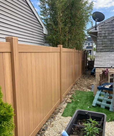 Tall wooden privacy fence beside a house, with a narrow gravel path and yard items along the side.