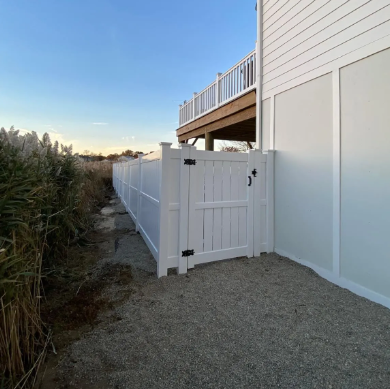 White side gate beside a house and deck, opening to a gravel path with tall shrubs and a white fence