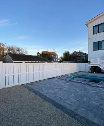 Backyard with white fence, paved patio, and swimming pool beside a house under a clear blue sky