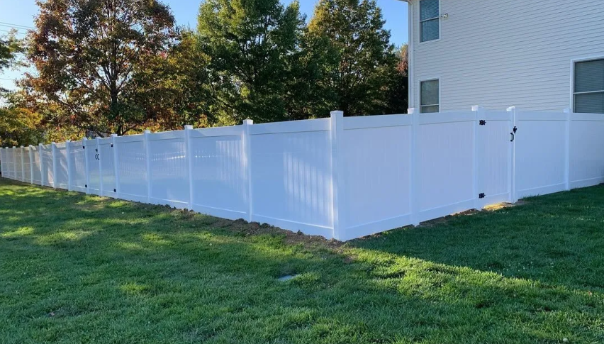 White vinyl fence surrounding a grassy yard beside a white house, with trees in the background