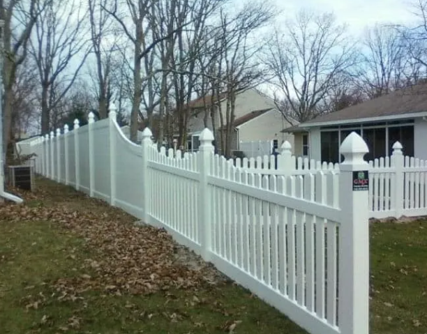 White vinyl fence with decorative posts along a sloped yard beside a house and bare trees