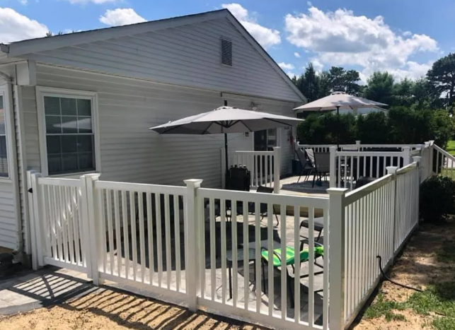 White fenced patio deck beside a gray house with umbrellas and outdoor seating on a sunny day