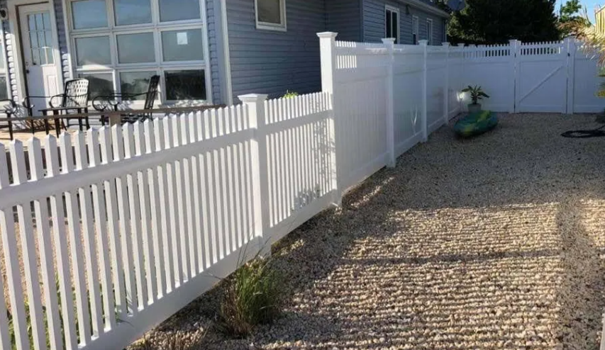 White picket fence beside a house, bordering a gravel path and patio area in bright sunlight