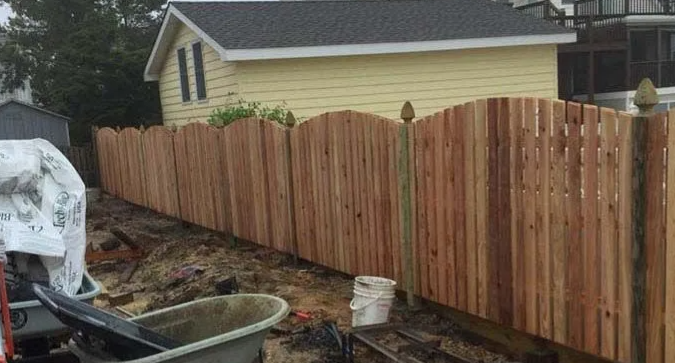 Freshly installed wooden privacy fence along a muddy yard beside a yellow house