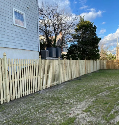 Backyard with a beige wooden fence beside a house, grass yard, and trees under a blue sky