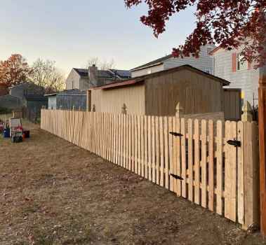 Wooden picket fence beside a tan shed in a suburban backyard at sunset
