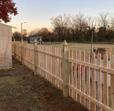 White picket fence along a grassy path at sunset, with trees and a playground in the distance