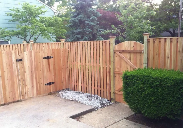 Wooden backyard gate with black hinges beside a trimmed hedge and concrete patio.