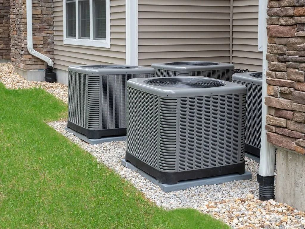 Four gray air conditioning units next to a building on a gravel bed, adjacent to grass.