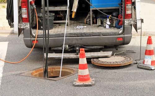 Utility truck with open rear near manhole covers, hoses, and orange traffic cones on a wet street.