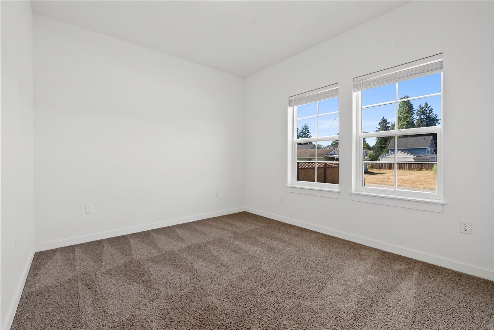 An empty bedroom with two windows and a brown carpet.
