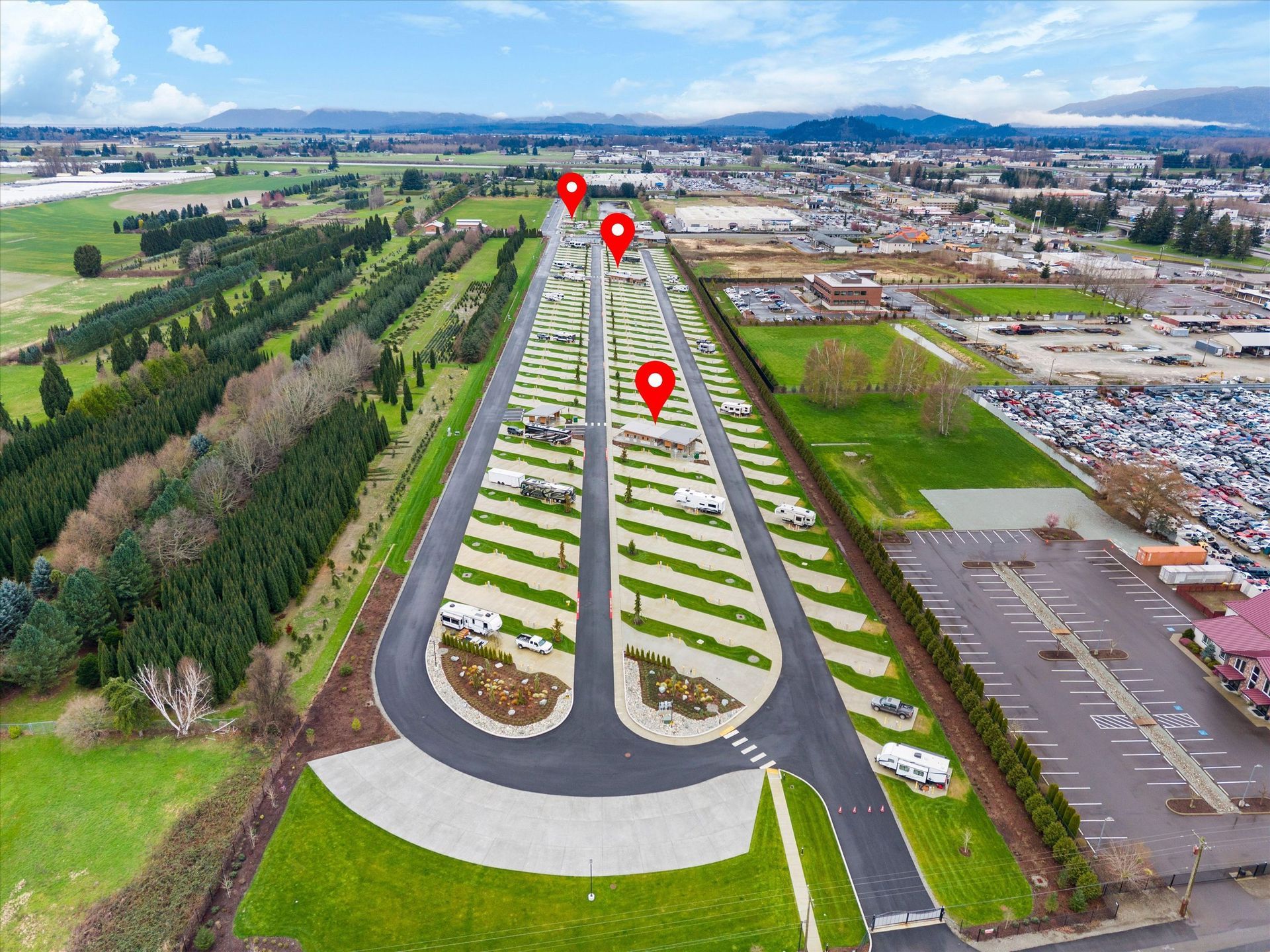 Aerial view of a caravan park with long, parallel rows of parking spaces, green grass, and three red location pins.