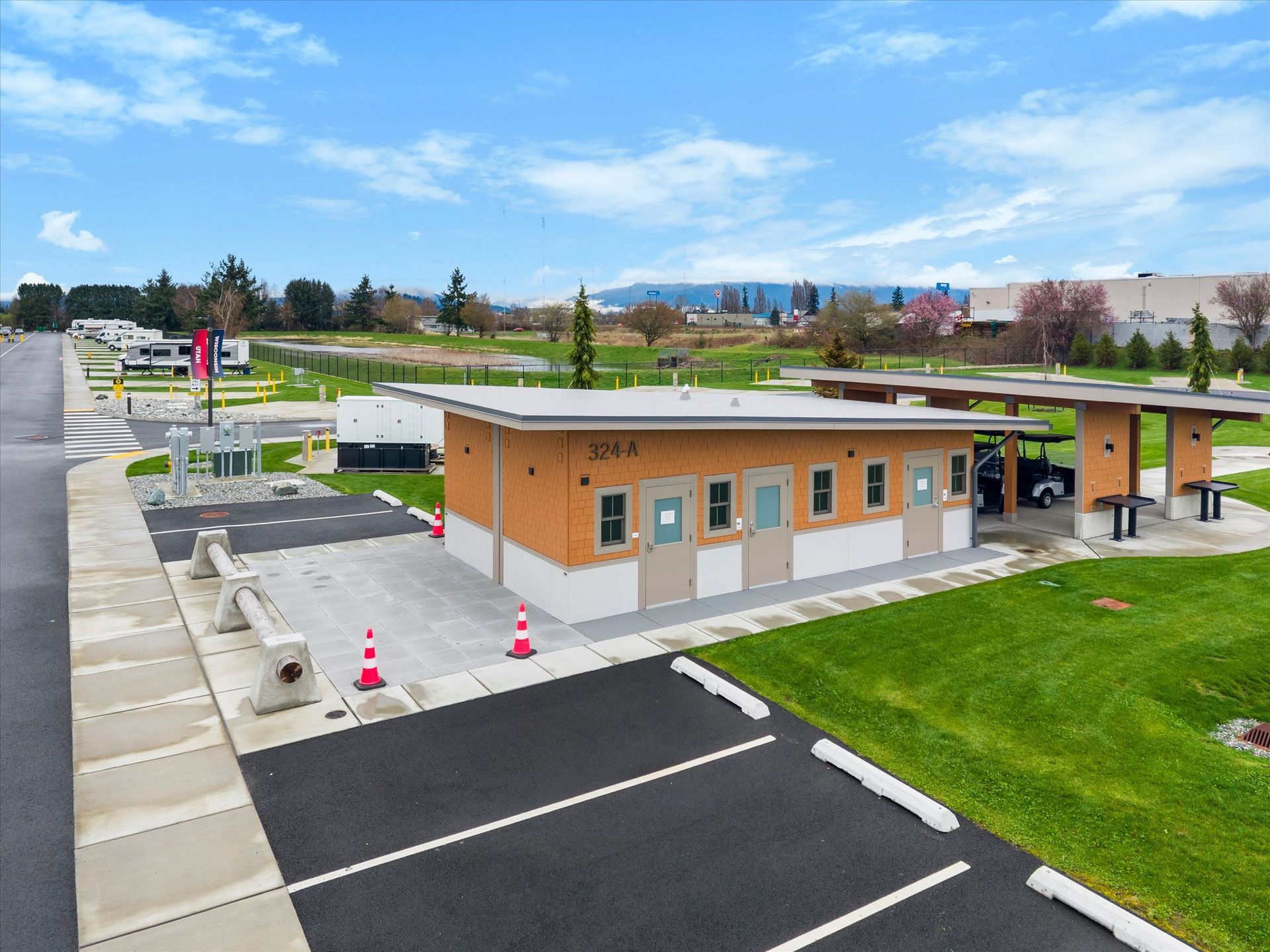 An elevated view of a tan and white restroom building with a slanted metal roof and parking spaces in a grassy park setting.