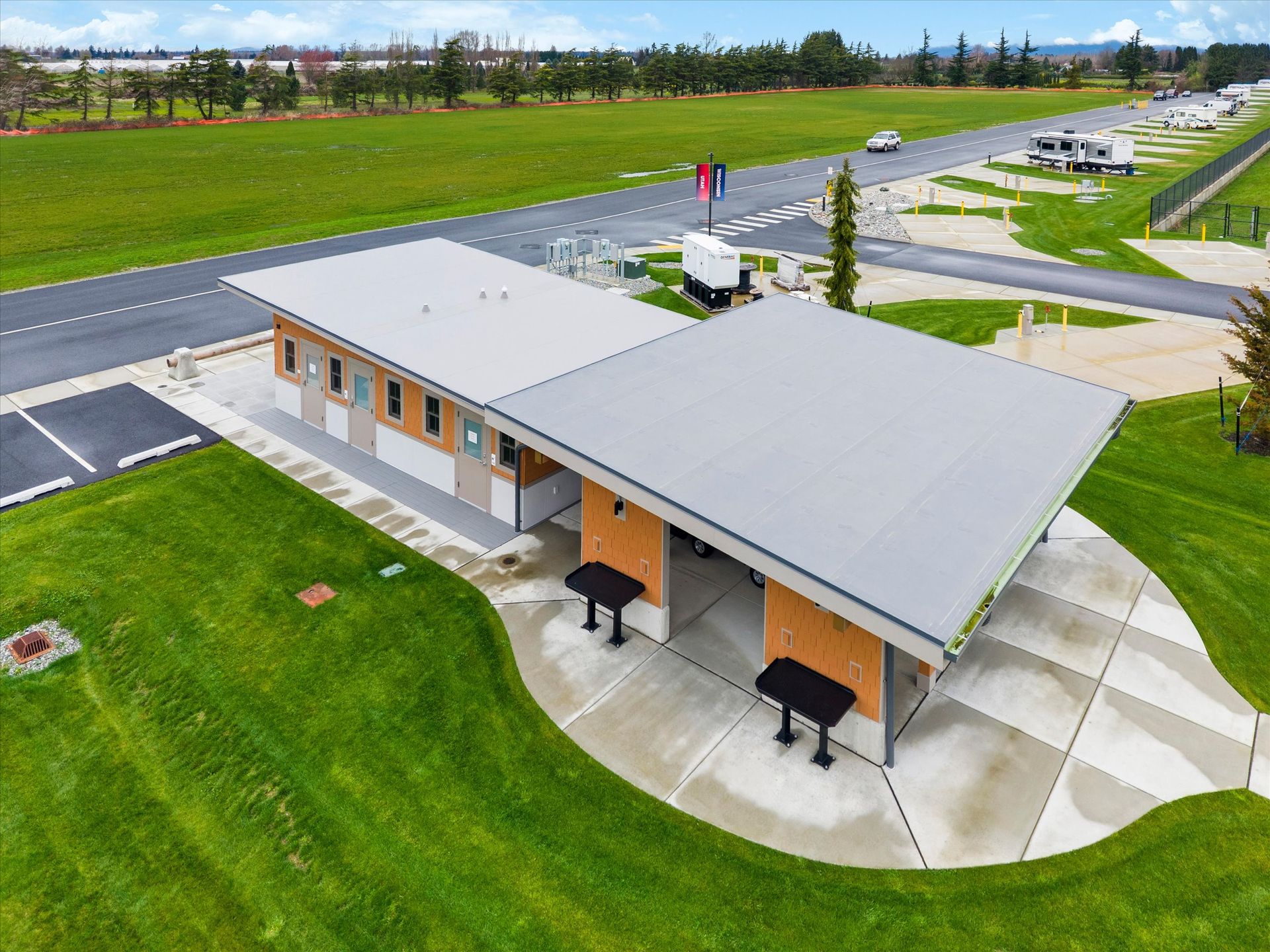 An aerial view of a modern public restroom and covered outdoor seating area with grills, set in a grassy park by a road.