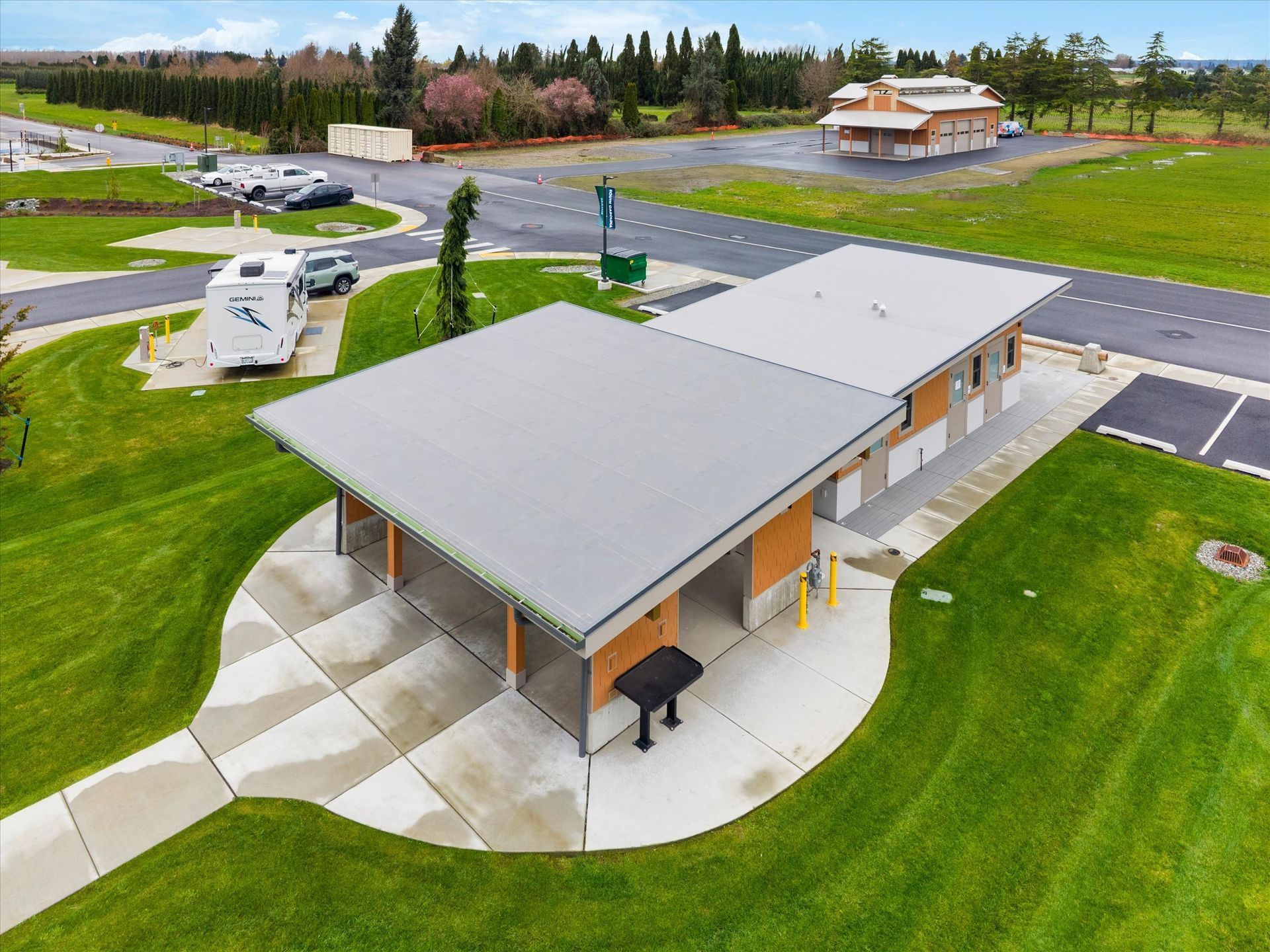 An aerial view of a modern public rest stop facility with a concrete pad, metal roof, and adjacent parking in a park.