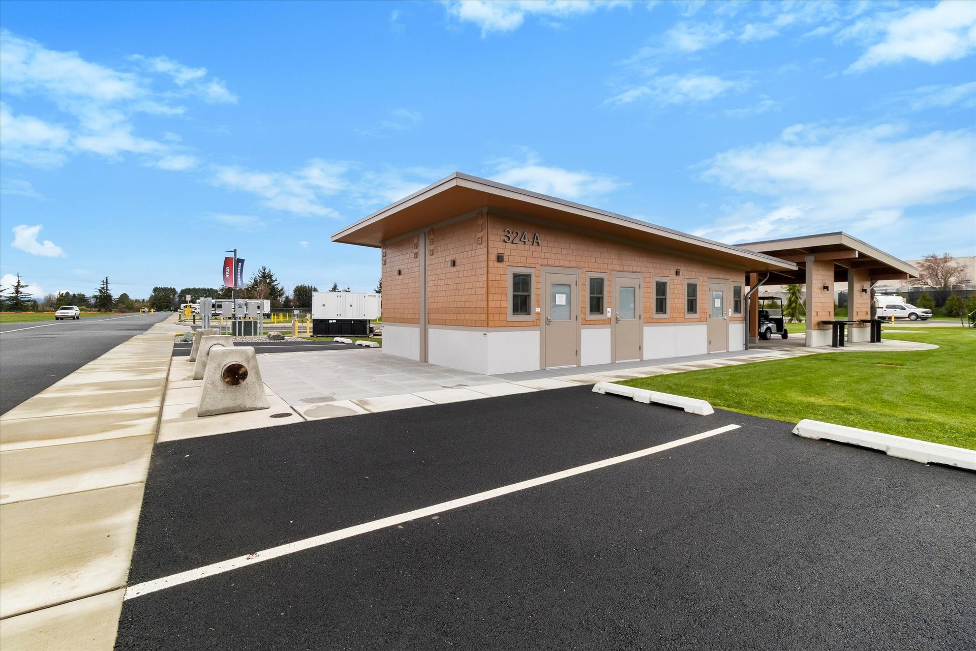 A single-story, wood-sided public restroom facility with an attached covered picnic area next to a paved parking lot.