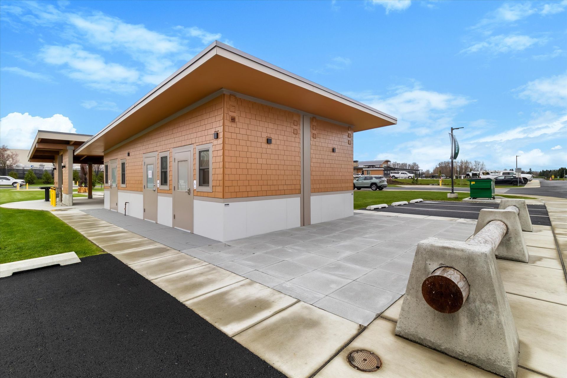 A modern, single-story public restroom building with tan siding and white trim located in an outdoor parking area.