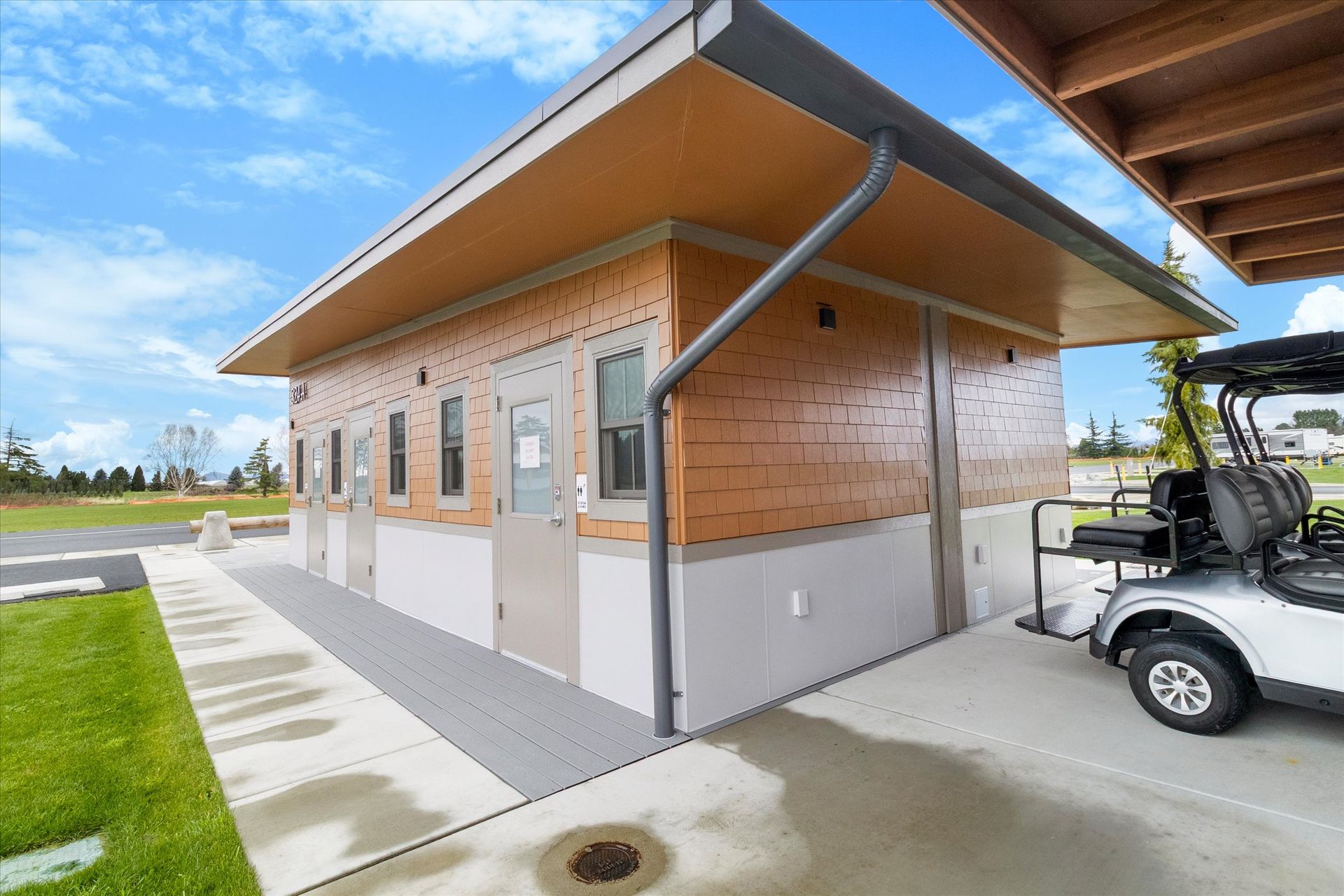 A modern, single-story public restroom building with light-brown siding, a grey concrete base, and a golf cart parked nearby.