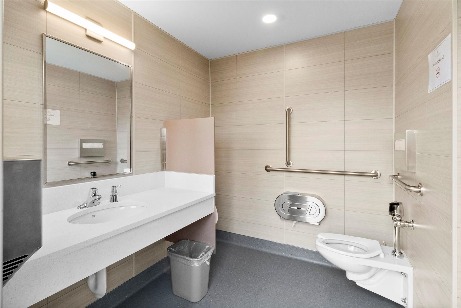 Accessible restroom with a white vanity sink, a wall-mounted toilet with grab bars, beige tile walls, and gray flooring.