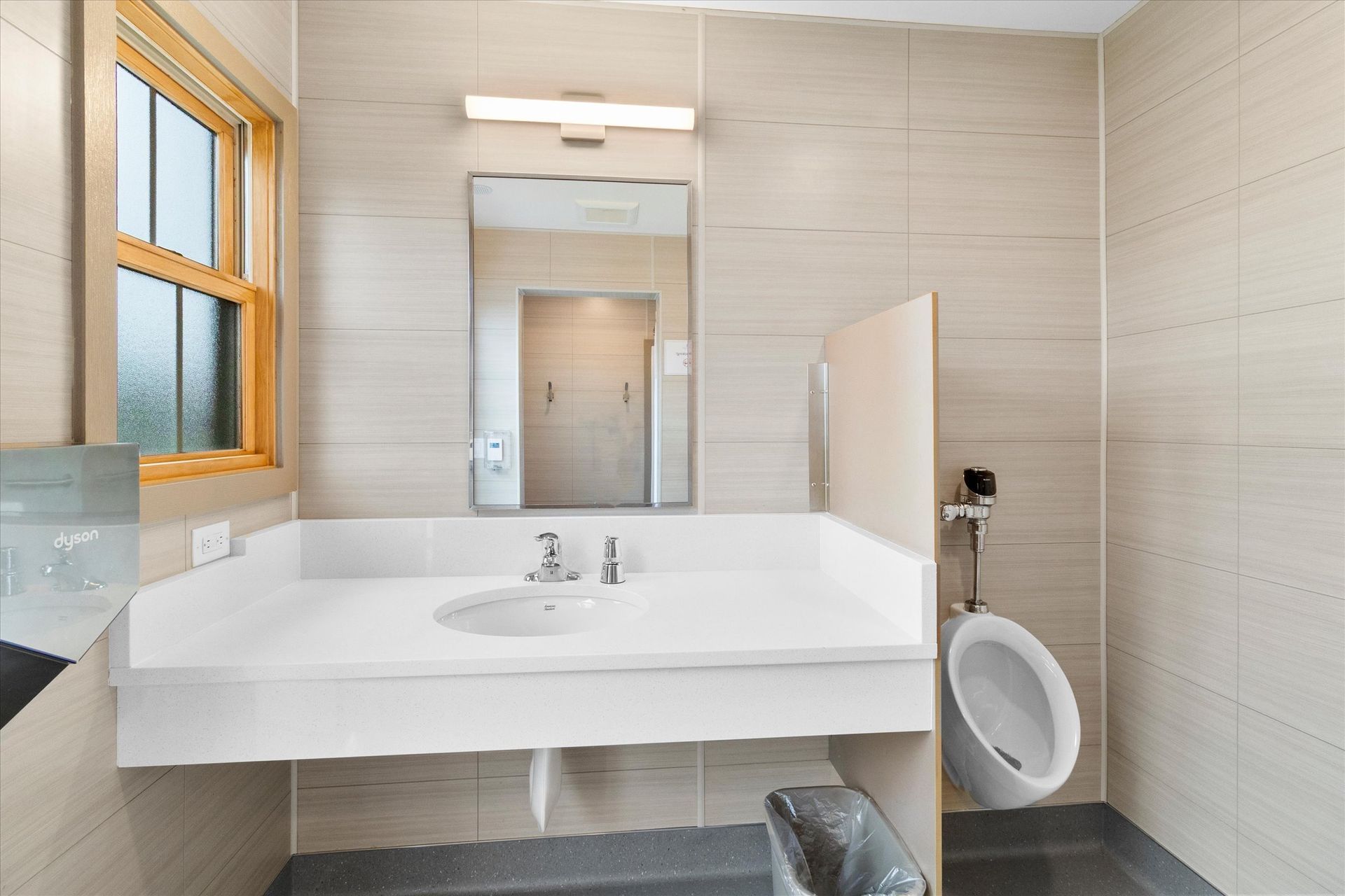 A bathroom with a wall-mounted white vanity sink, a mirror, and a urinal separated by a privacy partition.