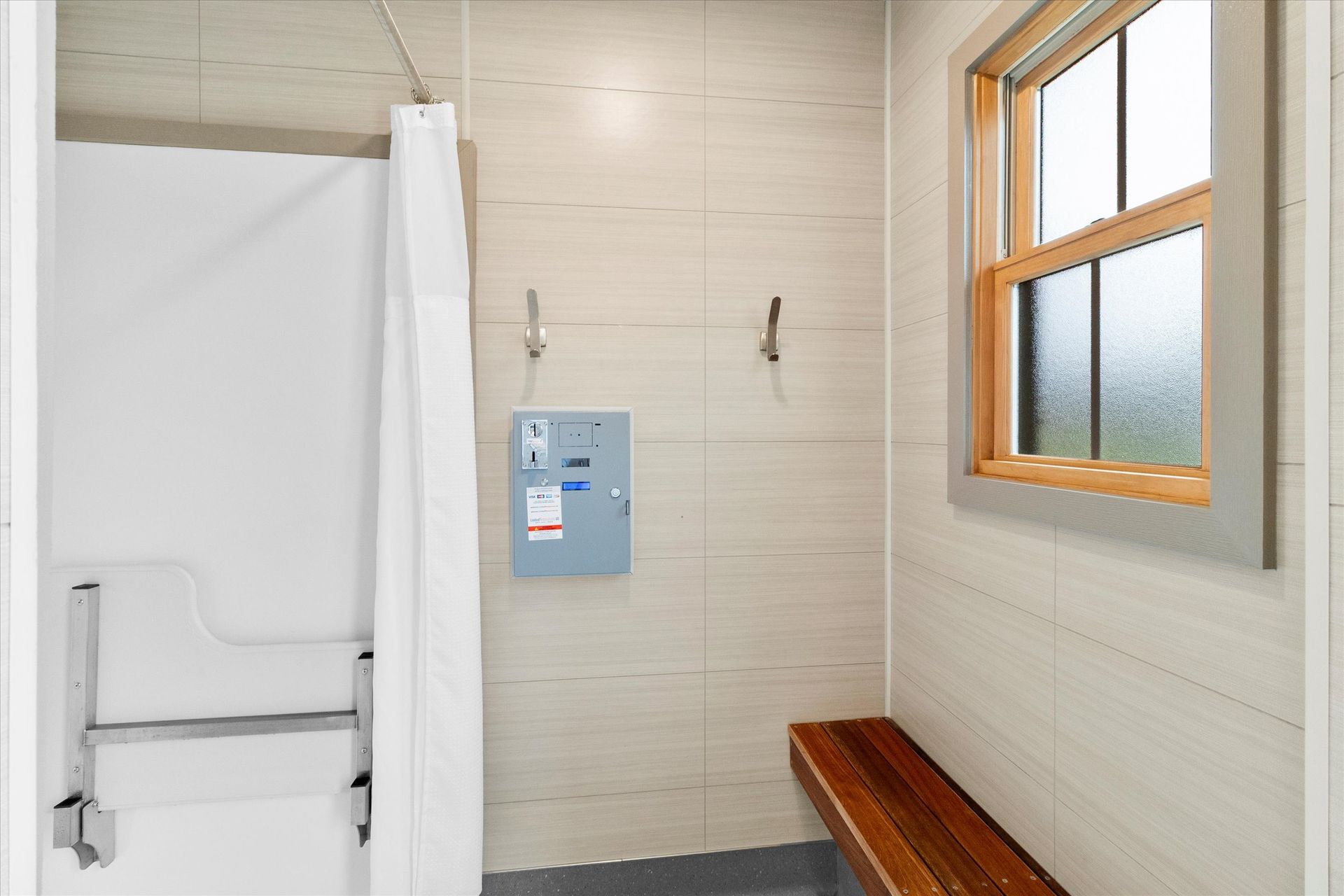 Accessible shower area with a fold-down bench, grab bars, a window, and an electrical panel on a beige tiled wall.