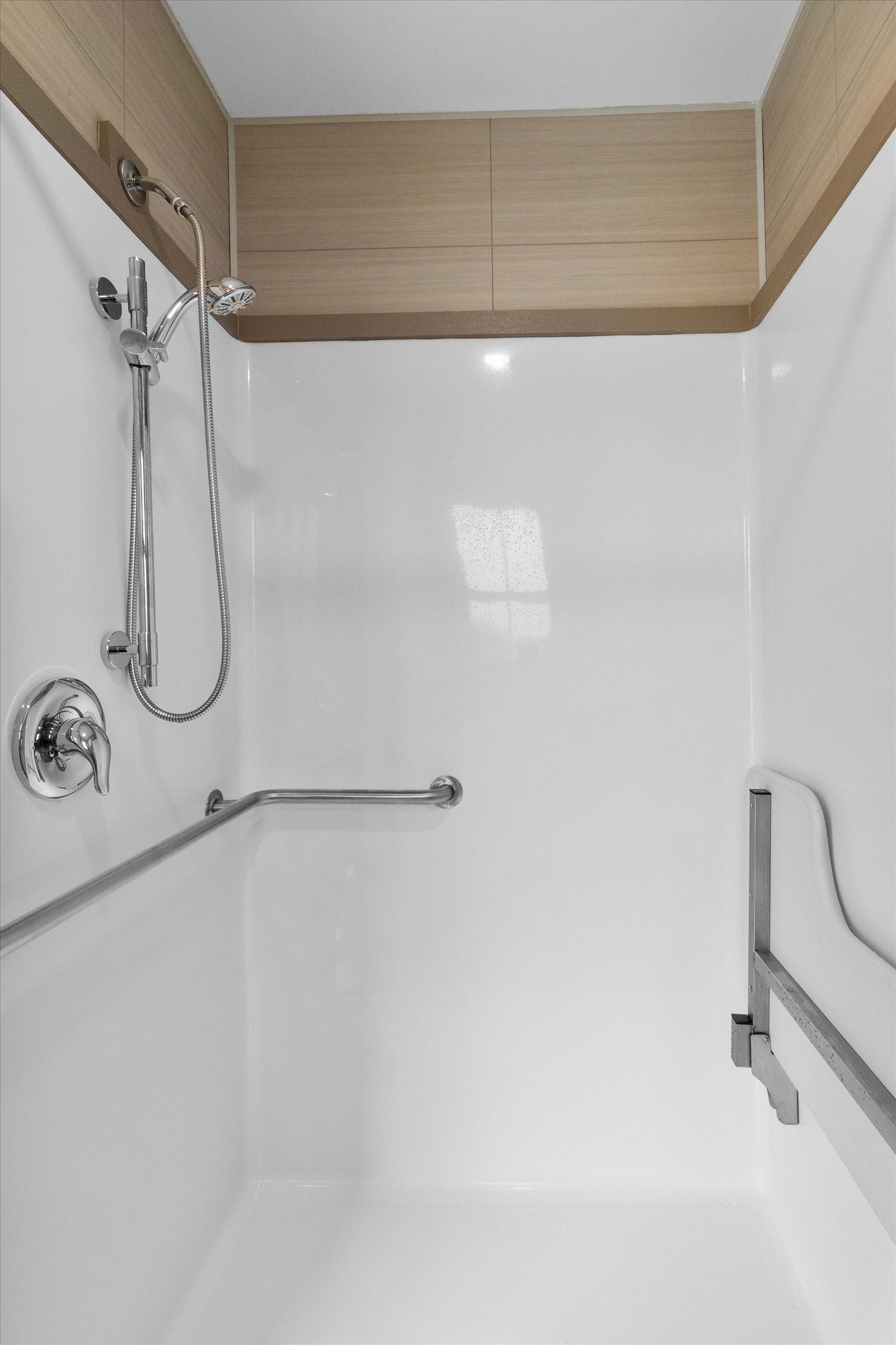 A modern shower with white walls, a chrome handheld shower head, and silver grab bars installed for accessibility.