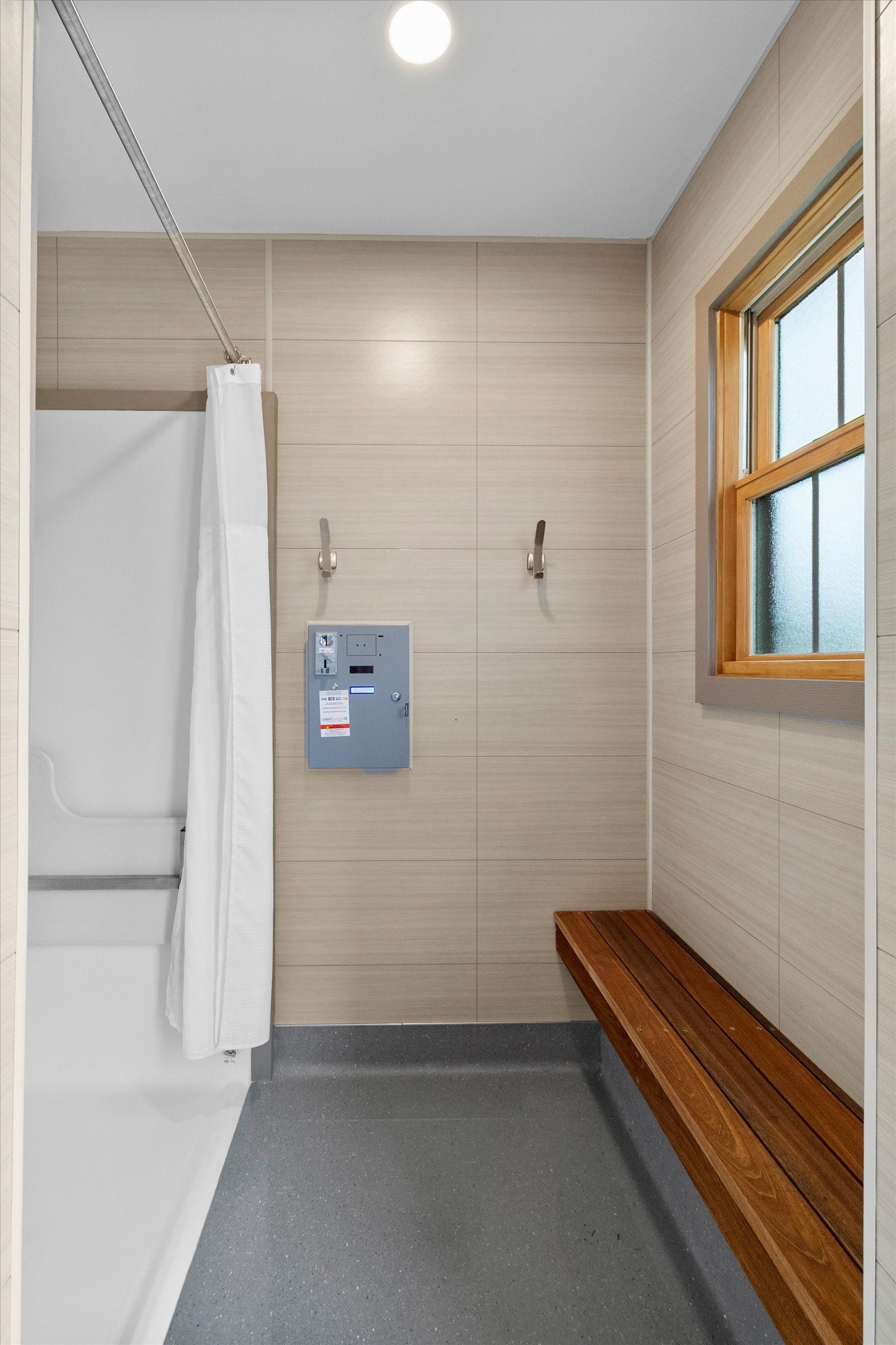 Accessible shower room with a walk-in shower, wooden bench, light-colored tiled walls, and a wall-mounted control unit.