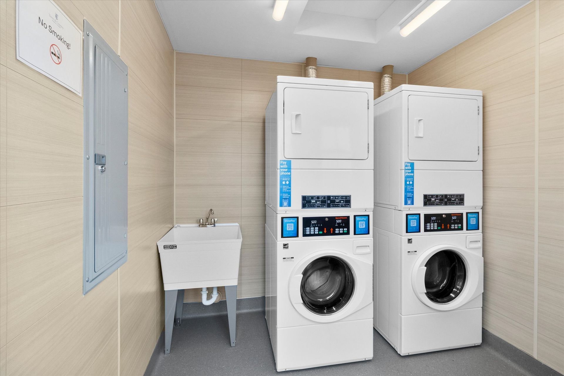 A laundry room with two white stacked washer-dryer units next to a utility sink against beige tiled walls.