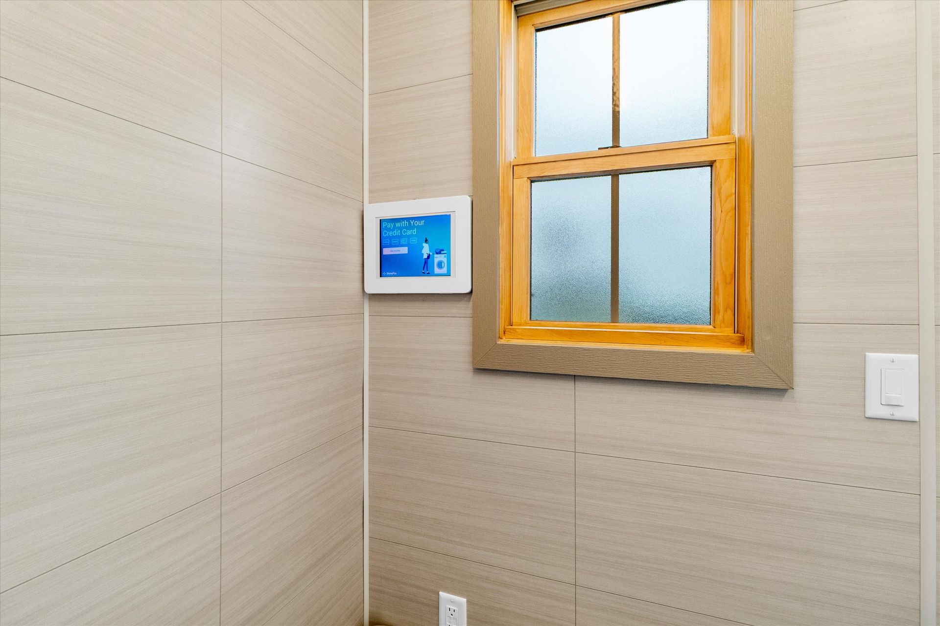 A beige-tiled wall with a wood-framed window, a small digital smart home controller, and a light switch.