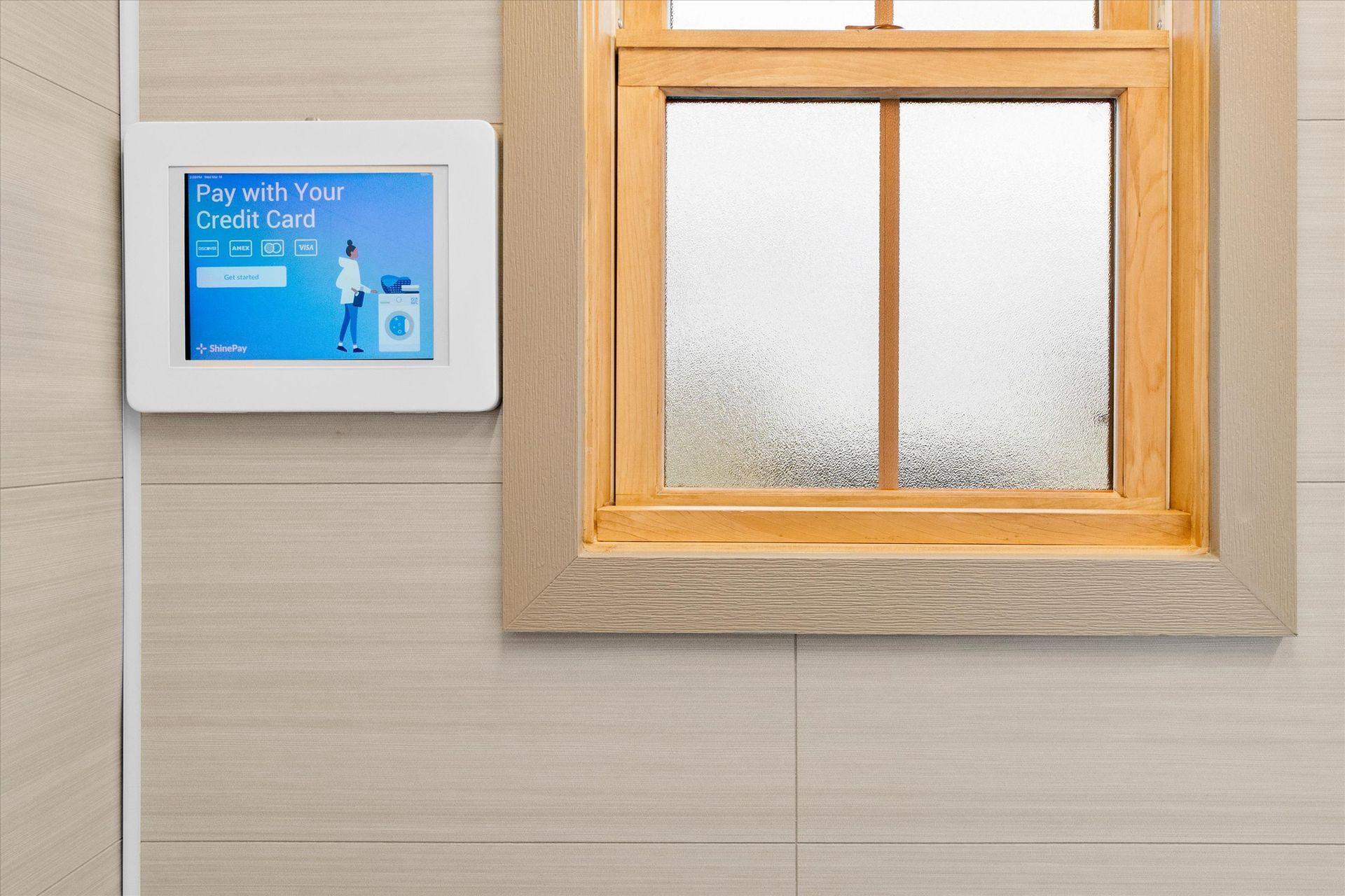 A digital payment screen mounted on a tiled wall next to a wooden-framed window, displaying a credit card payment prompt.