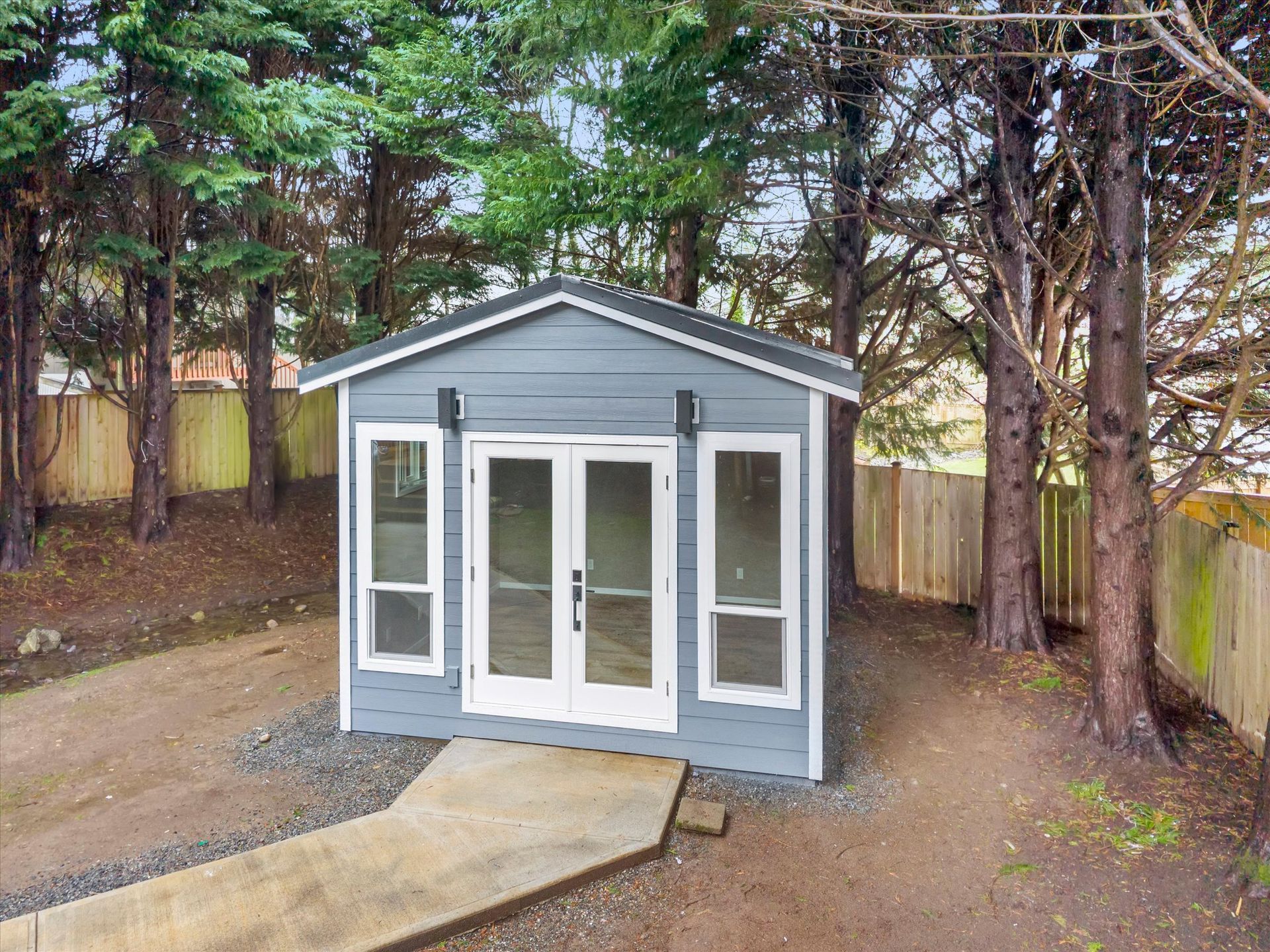 A small, grey backyard studio with double glass doors and two tall windows, set in a wooded area with a concrete path.