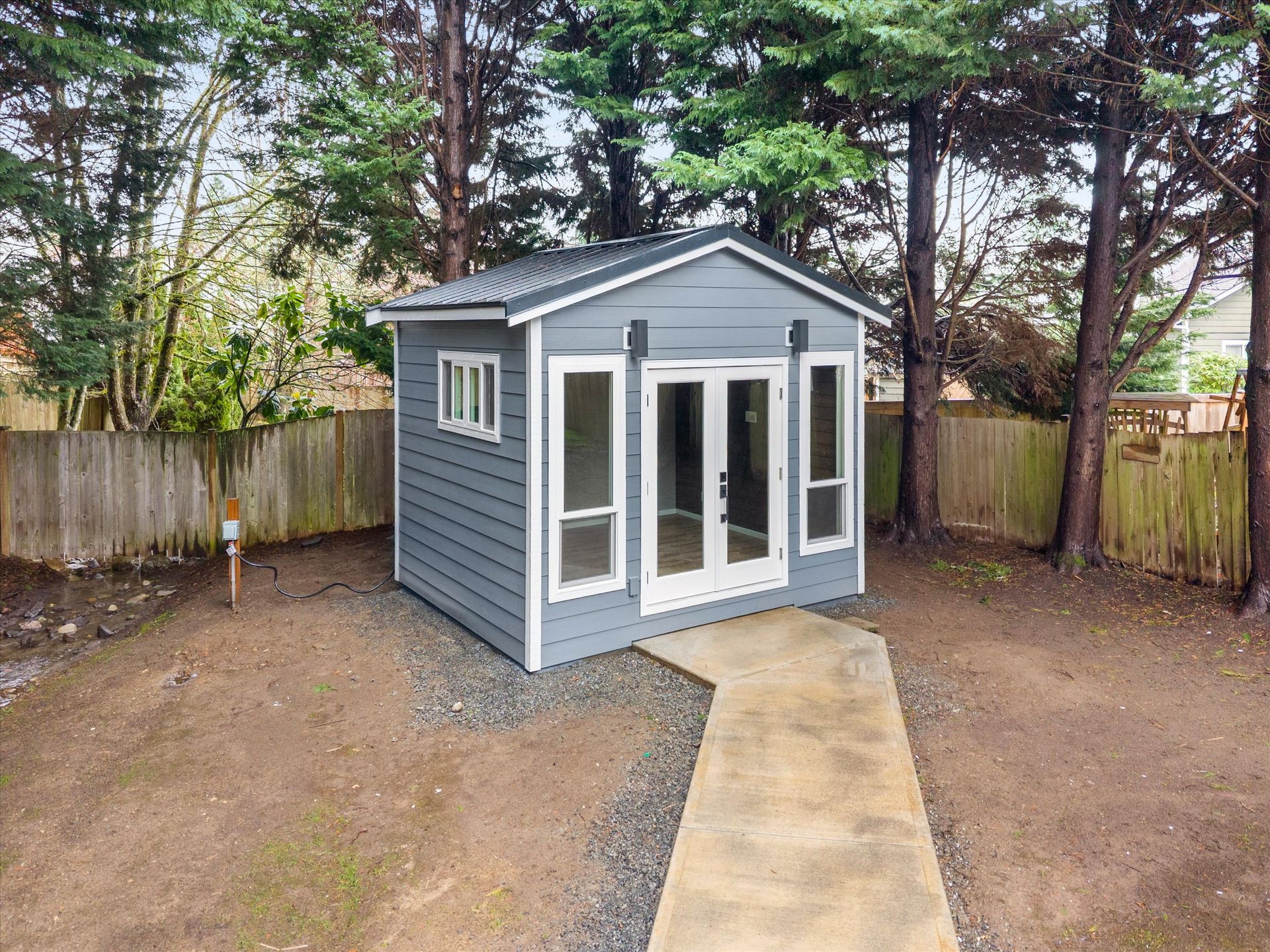 A grey shed with white trim, French doors, and windows, set in a backyard with a concrete path leading to the entrance.