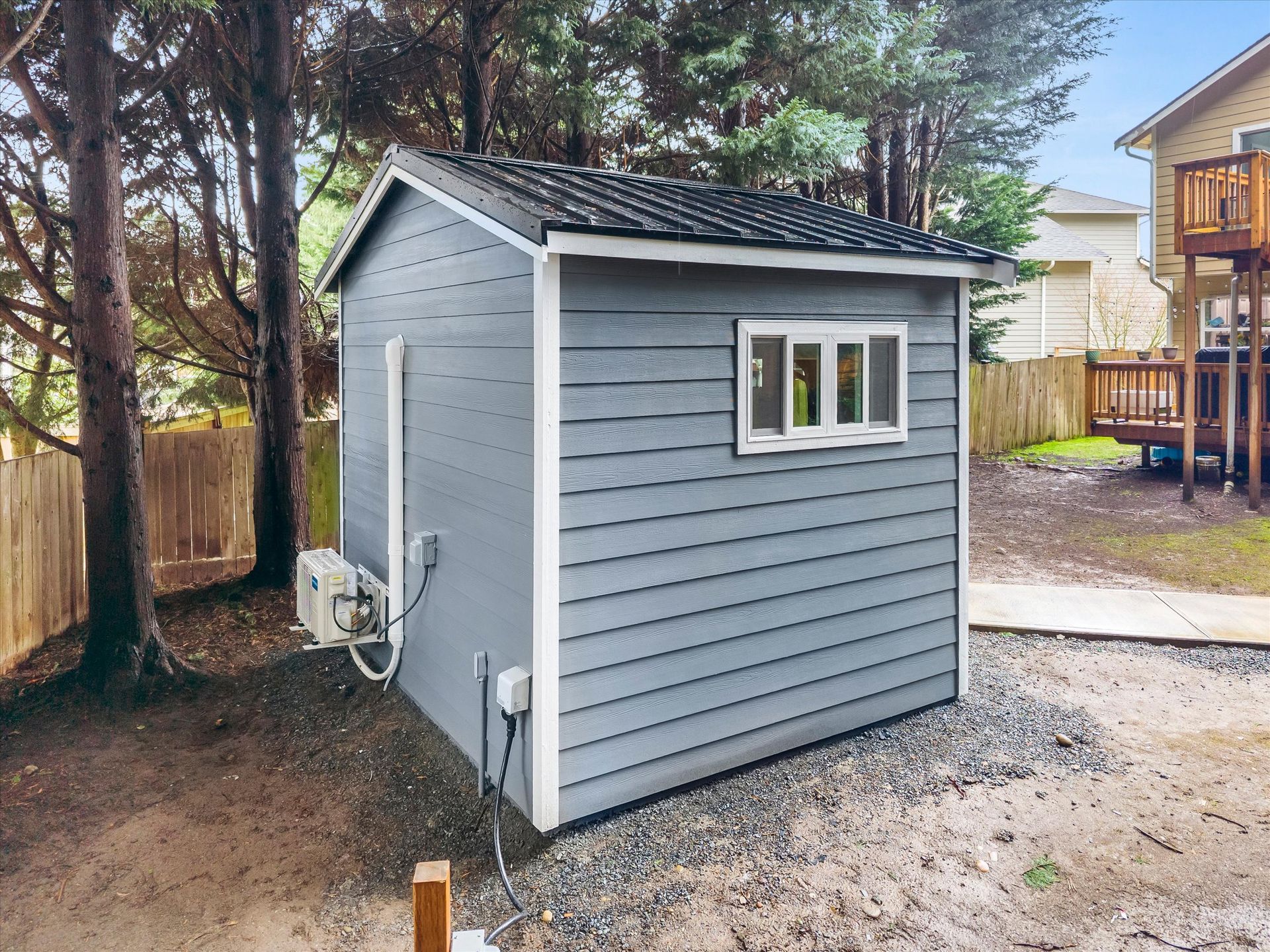 A gray, rectangular backyard shed with a black roof, a small window, and an external HVAC unit, situated in a wooded area.