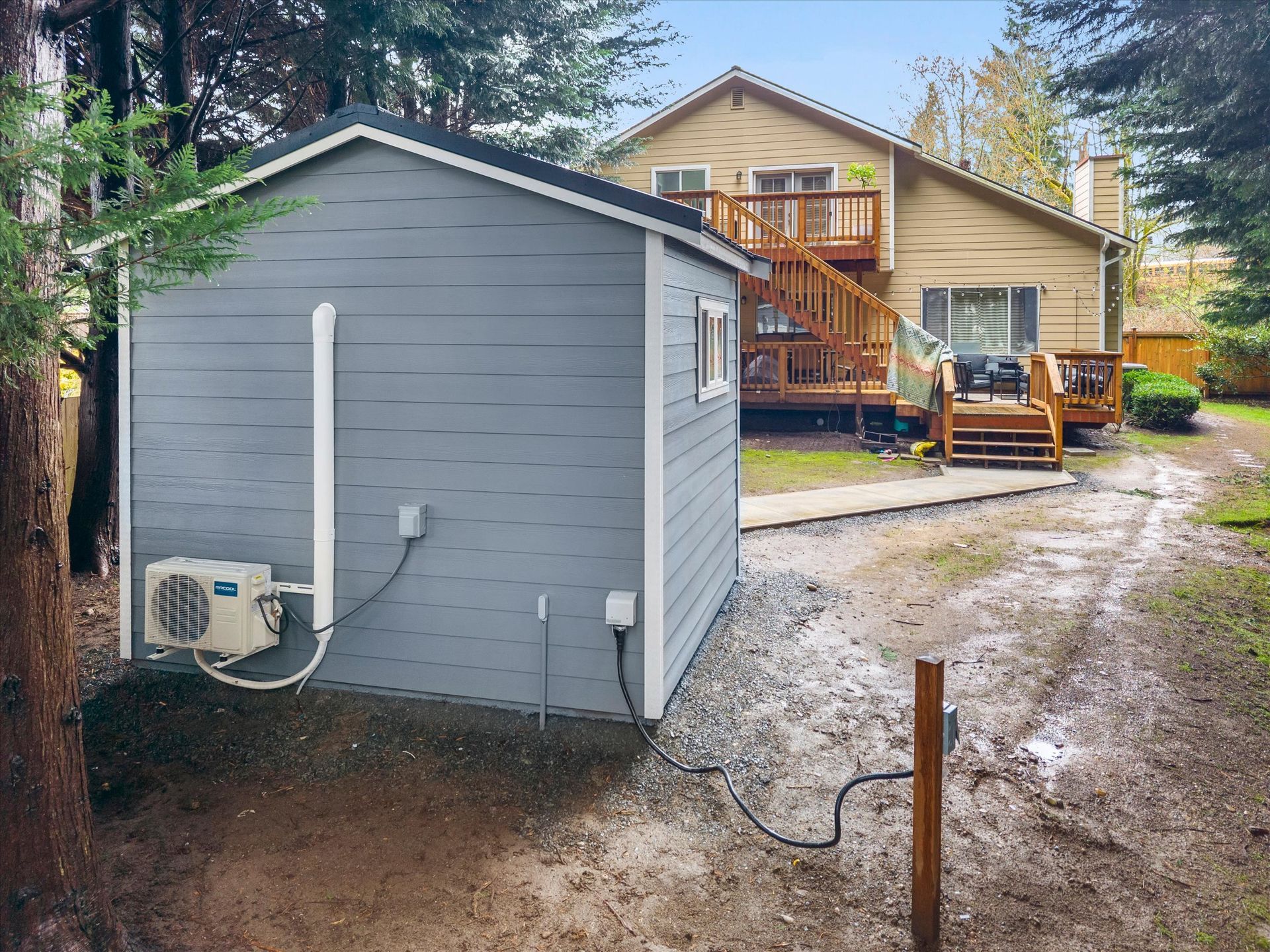 A gray backyard shed with an exterior HVAC unit sits in a dirt yard near a two-story house with a wooden deck.