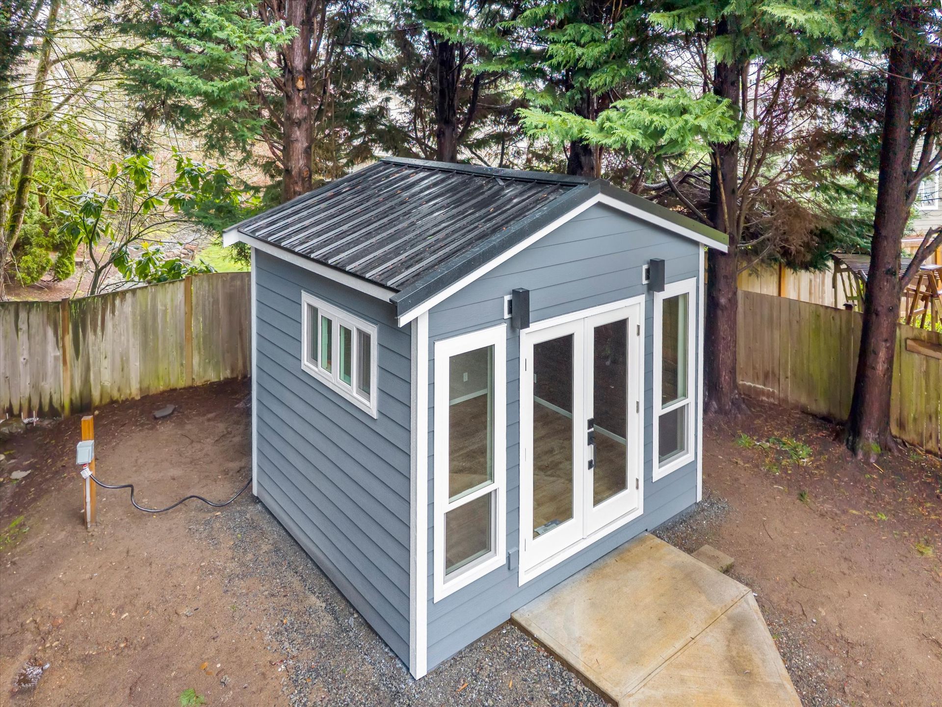 A blue, modern shed with glass double doors and windows, situated in a wooded backyard on a concrete pad.