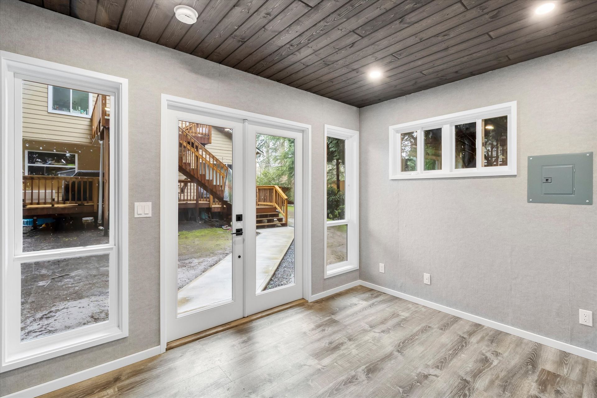 A bright, empty room with light gray walls, wood-look flooring, a dark plank ceiling, glass doors, and large windows.